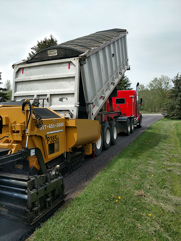 Red dump truck feeding yellow LeeBoy paver on a rural drive